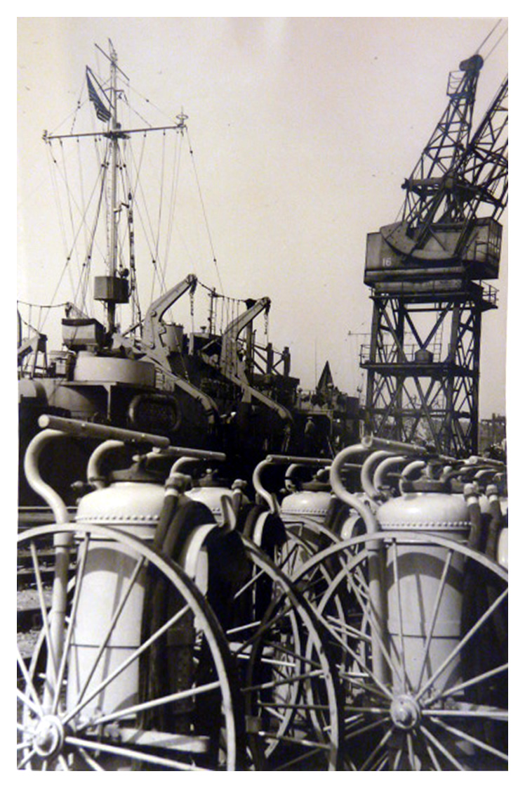A photograph of US naval ships moored at Penarth Dock with what appears to be fire extinguishers lined up along the quay. The photograph was most probably taken between November 1943 and June 1944. 